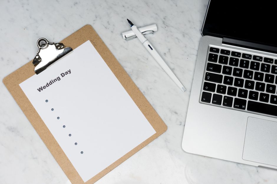 A close-up view of a desktop workspace featuring a brown clipboard with a white paper titled 'Wedding Day' and bullet points, a white ballpoint pen, and a partially visible open laptop with a black ke