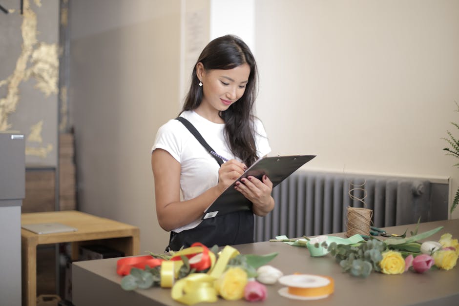A young woman with long dark hair, wearing a white t-shirt and black apron, is standing at a worktable in a floral workshop, writing on a clipboard. The table in front of her is arranged with fresh fl