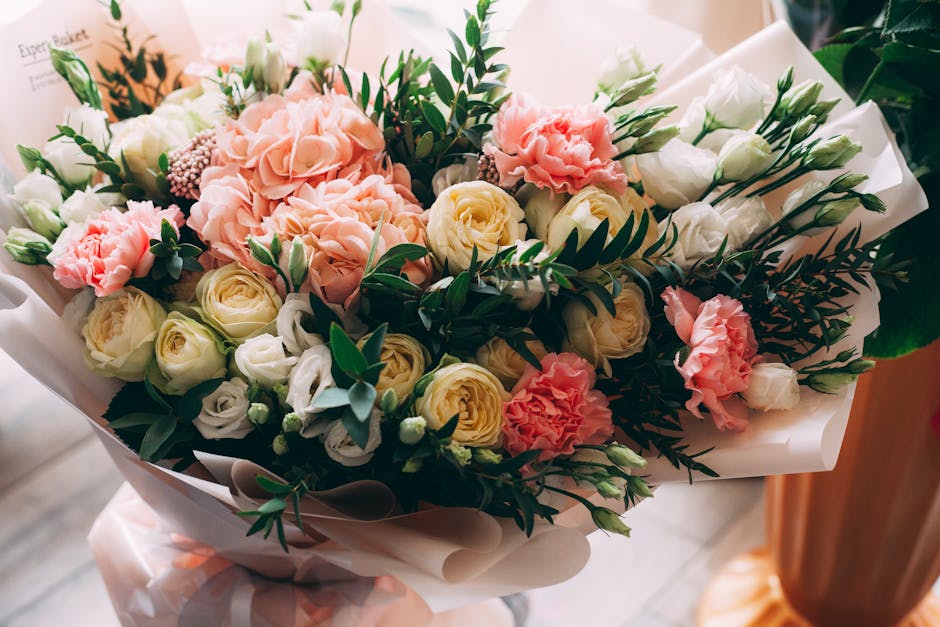 A bouquet of fresh flowers featuring soft pink carnations, creamy white roses, and pale yellow ranunculus, accented with dark green fern and foliage, arranged in a compact, rounded style. The bouquet 