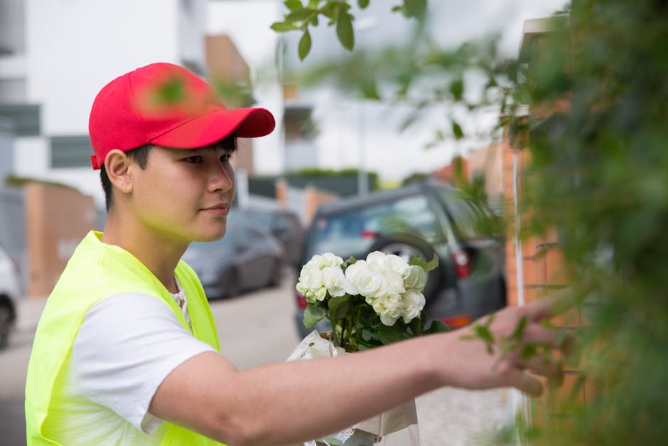 A young male flower delivery courier, dressed in a high-visibility yellow vest and a red cap, is delivering a floral gift outside a residential building. He is holding a bouquet of fresh white hydrang